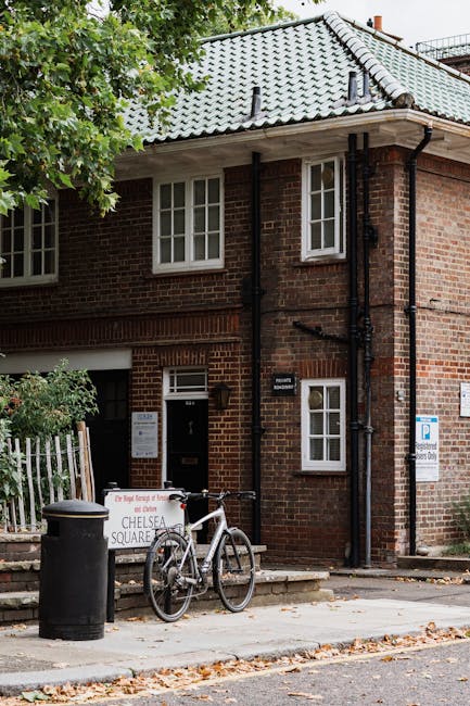 The exterior of a brick building in Sloane Square Chelsea is shown with a grey tiled sloped roof and white-framed sash windows. In front, there is a paved sidewalk with a bicycle parked against a blac